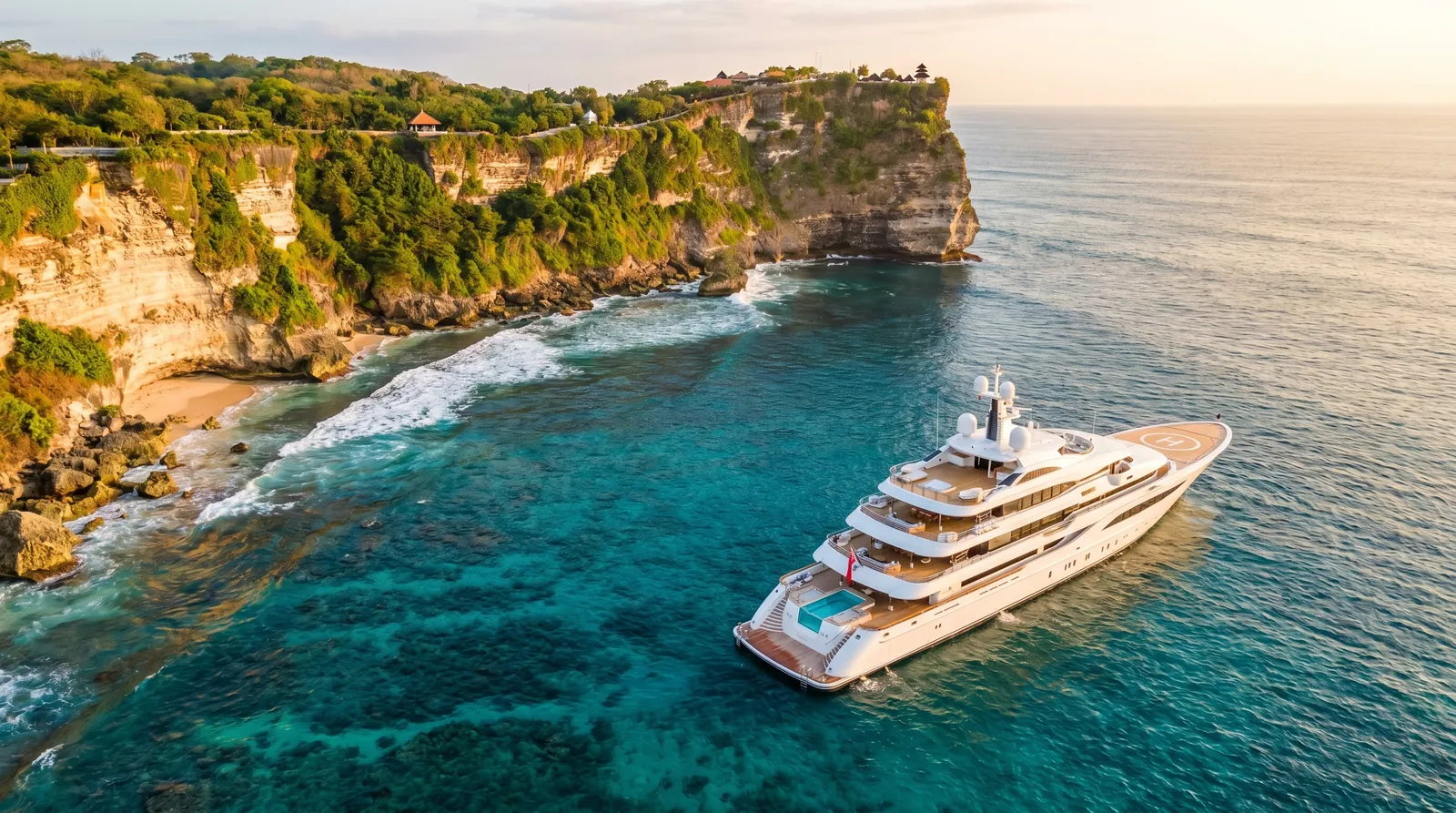 Aerial view of a luxury yacht in turquoise water beside a dramatic Bali cliff coastline at golden hour